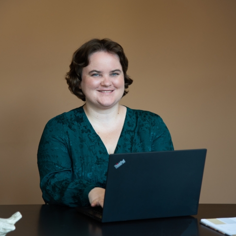 Photo of Katherine wearing a green dress sitting at a desk and working on a laptop computer.