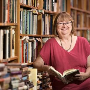 Woman holding book open leaning against a bookshelf, filled with many colourful books.