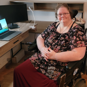 Photo of Rosella Derksen seated in a wheelchair with an office desk and her laptop computer and external monitor in the background.